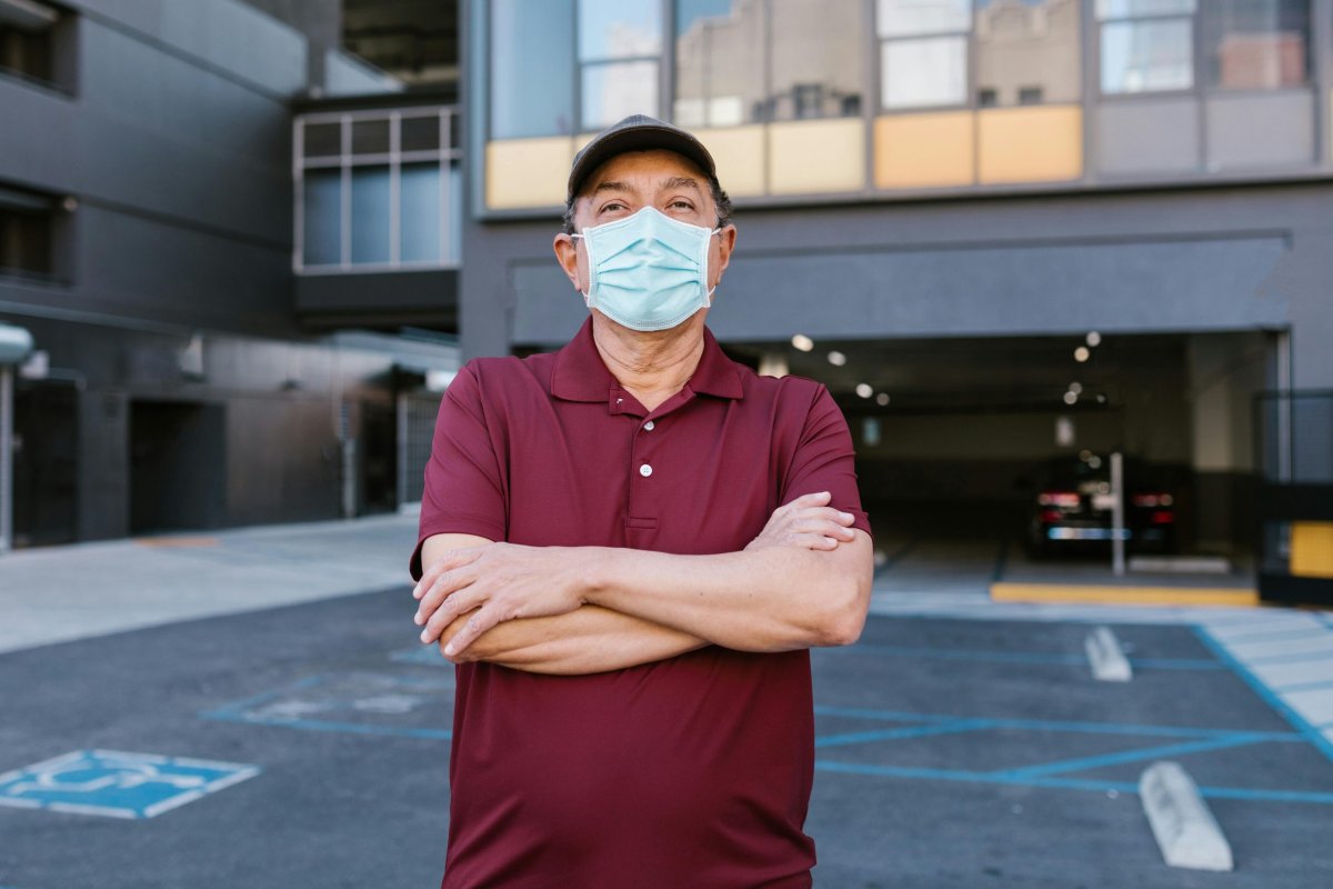 An older man wearing a mask, standing outside with his hands crossed