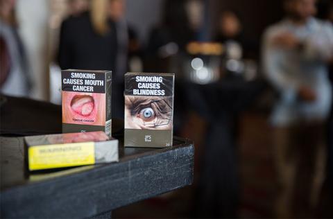 Plain packaging in Australia - tobacco packages in drab dark brown packaging with large graphic health warnings, one with a picture of a tumor and text saying Smoking Causes Mouth Cancer, another with a picture of a damaged eye saying Smoking causes blindness